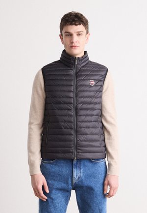 Gilet imbottito nero con colletto alto, chiusura con zip e patch con logo. Indossato sopra una maglietta a maniche lunghe beige, abbinato a jeans blu.