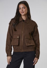Brązowa kurtka bomber wykonana z syntetycznego zamszu, z zapięciem na zamek, naszytymi naramiennikami, dwiema przednimi kieszeniami i elastycznym pasem. Gładka tekstura.