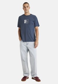 Navy blue cotton t-shirt with a white graphic print on the chest, paired with light blue denim pants and burgundy footwear.