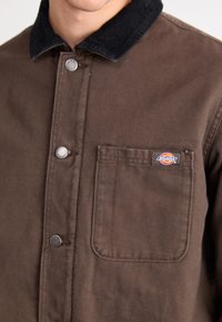 Brun canvasjacka med svart kordflöjelskrage, tryckknappar och en bröstficka med en Dickies-logotyp. Slät textur.