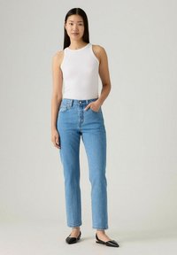 Top de tirantes blanco acanalado, vaqueros de tiro alto en un azul claro con un corte relajado y zapatos planos negros; el modelo está de pie con una mano en el bolsillo.