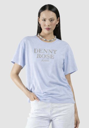 Donna con capelli lunghi e scuri che indossa orecchini a cerchio, una collana di perle, una maglietta azzurra "DENNY ROSE JEANS" e pantaloni bianchi, mano in tasca.
