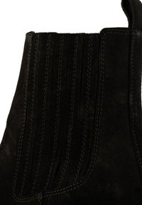 Botín de tobillo de ante negro con líneas de costura verticales, una textura suave y una lengüeta en la parte posterior para facilitar su uso.