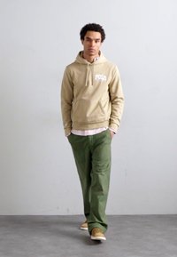 Sweat à capuche beige avec un logo blanc, pantalon vert olive et baskets beiges. Tenue décontractée avec des couleurs distinctes et une coupe confortable.