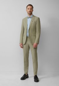Costume vert clair à coupe ajustée, associé à une chemise bleu clair à boutons et un nœud papillon gris. Des chaussures noires complètent le look.