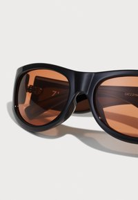 Gafas de sol negras de gran tamaño con lentes redondos, tintado marrón y montura de plástico suave. Detalles notables incluyen brazos gruesos y una sutil marca.