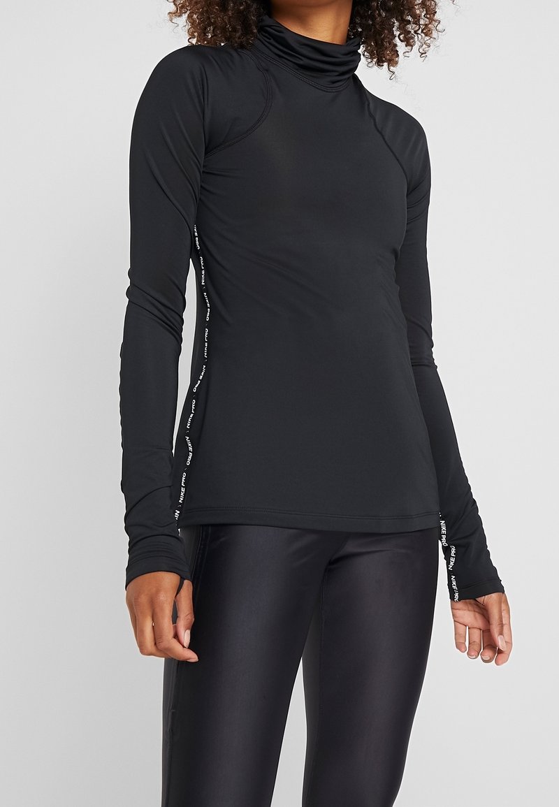 Schwarzes Langarmoberteil mit hohem Kragen. Verfügt über eine schmale Passform, weiche Textur und grafische Akzente entlang der seitlichen Nähte. Kombiniert mit glänzenden schwarzen Leggings.