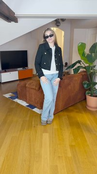 Svart cropped jakke med gullknapper, hvit skjorte, lyseblå jeans og grå støvler; innendørs setting med en myk brun sofa og grønn planter.