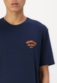 Námornícky modré tričko s krátkym rukávom vyrobené z bavlny. Na hornej ľavej časti hrudníka sa nachádza oranžová vyšitá značka "WRANGLER JEANS 1947".