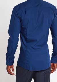 Marineblaue Langarmbluse mit abgerundetem Saum, taillierter Passform und geknöpften Manschetten. Glatter Stoff mit minimaler Verzierung und ohne Muster.