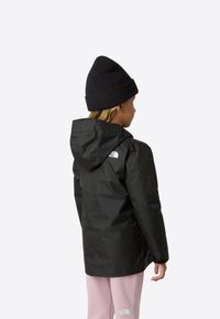 Černá voděodolná bunda s kapucí, se bílým logem na rameni, v kombinaci s černou pletenou čepicí a růžovými kalhotami.