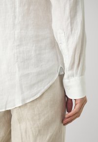 Camisa de lino blanca con mangas largas, puños abotonados y dobladillo curvado. Etiqueta visible en la costura lateral. Textura ligera y transpirable.