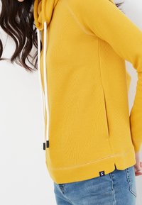 Gul huvtröja med ribbad textur, vita dragsko och sidoslitsar. Har en liten logotyp vid fållen.