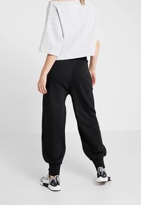 Persoon in zwarte joggingsbroek, grijs cropped T-shirt en witte Puma sneakers, staand met de rug naar de camera tegen een effen witte achtergrond.