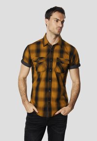 Chemise à manches courtes à carreaux jaune moutarde et noir. Dispose de deux poches poitrine, d'une fermeture boutonnée et de manches retroussées.