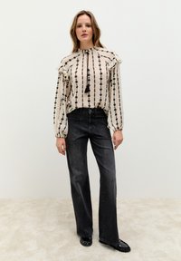 Hellfarbene, gemusterte Bluse mit Rüschenschultern und einer Schnürung am Ausschnitt, kombiniert mit dunklen Schlagjeans und schwarzen flachen Schuhen.