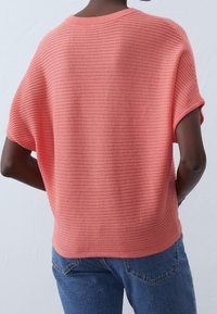 Korálový pletený top s ribovanou texturou, krátkými rukávy a volným střihem, kombinovaný s modrými džínami.