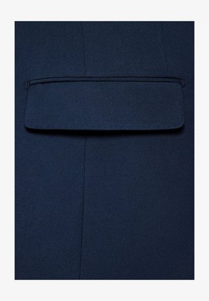 Granatowy materiał o fakturowanej powierzchni z prostokątną kieszenią na górze. Widoczne szwy podkreślają strukturę.