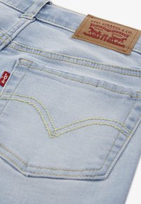 Ανοιχτό μπλε τζιν με καφέ δερμάτινο σημάδι στη μέση, κίτρινη ραφή και κόκκινη ετικέτα με λογότυπο στην πίσω τσέπη.