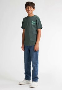 Vihreä puuvillainen t-paita, jossa on pyöreä pääntie ja rintakehällä graafinen painatus. Yhdistetty sinisiin farkkuihin ja valkoisiin lenkkareihin.