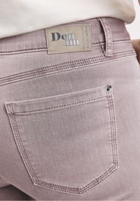 Los jeans de mezclilla de color rosa claro presentan un bolsillo trasero, costuras reforzadas y una etiqueta color beige con "Den im Gerny Wieser". Textura suave, corte ajustado.