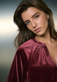 Haut en velours bordeaux avec un col plissé et une texture douce, présenté sur un fond flou et neutre. Un éclat subtil met en valeur la profondeur du tissu.