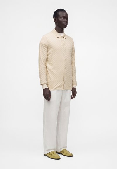 Homme portant une chemise beige à manches longues boutonnée, un pantalon blanc à jambes larges et des mocassins jaune clair, debout devant un fond blanc uni.
