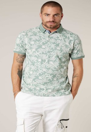 Homme aux cheveux courts et tatouages portant un polo à imprimé tropical vert et blanc ainsi qu'un pantalon cargo blanc, mains dans les poches, fond neutre.