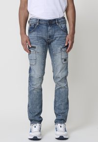 Pantalones cargo de denim azul claro con múltiples bolsillos, un corte ajustado, costuras visibles y un ligero efecto de lavado. Combinados con zapatillas blancas.