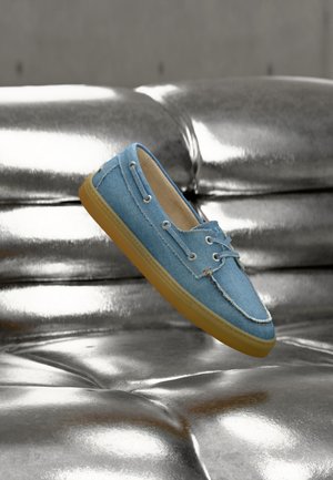 Chaussure décontractée en denim bleu avec doublure intérieure beige et semelle en gomme, flottant au-dessus d'une surface en cuir argenté capitonnée brillante.