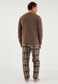 T-shirt à manches longues en polaire marron avec un col rond, associé à un pantalon de pyjama à carreaux dans des tons de marron et de blanc. Tissu doux, coupe décontractée.