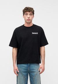 Νεαρός άνδρας που φοράει μαύρο t-shirt Timberland με το λογότ υπο και ανοιχτόχρωμα μπλε τζιν, στέκεται μπροστά σε απλό λευκό φόντο.