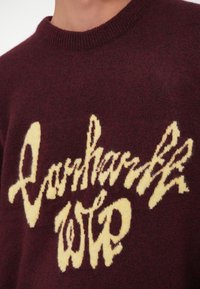 Burgundi kötött pulóver, melye előtérbe helyezkedik egy hangsúlyos sárga grafikai szöveggel. Puha tapintású, klasszikus kerek nyakú fazonnal.