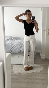 Juodas prigludęs viršus su V formos iškirpte, baltos platėjančios kelnės ir rudi šlepetės. Fone matyti lova su pilkai dryžuotais patalynės užvalkalais ir paprastu kilimu.