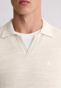 Helle beige gestrickte Poloshirt mit strukturiertem Finish und V-Ausschnitt. Dezentes Logo auf der Brust. Über einem weißen T-Shirt getragen.
