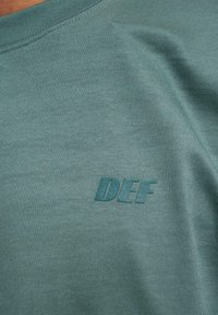 Teal short-sleeved shirt made of textured fabric, featuring a tonal "DEF" logo on the chest. Smooth finish with a crew neckline.