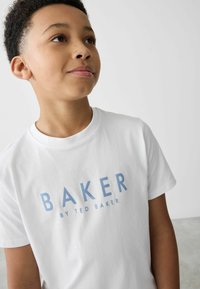 Bílý bavlněný tričko s modrým nápisem "BAKER" a "by TED BAKER", krátké rukávy, kulatý výstřih a jednoduchý design. Pozadí je neutrální.