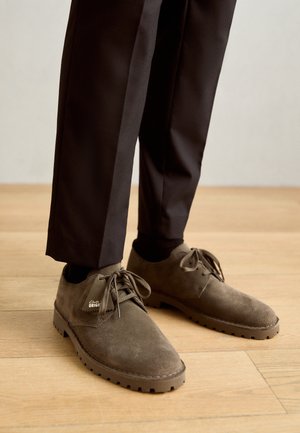 Personne portant un pantalon marron foncé et des chaussures en suède marron à lacets, debout sur un sol en bois clair.