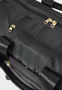 Fekete szövet táska dupla cipzárral, arany színű kiegészítőkkel, texturált pántokkal és több cipzáras zsebbel a rendezett tárolás érdekében.