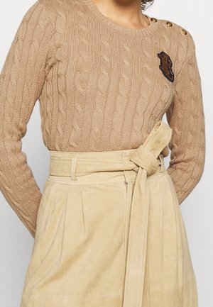 Pull beige en maille torsadée avec des boutons sur l'épaule et un patch brodé foncé, associé à une jupe beige taille haute avec ceinture nouée.