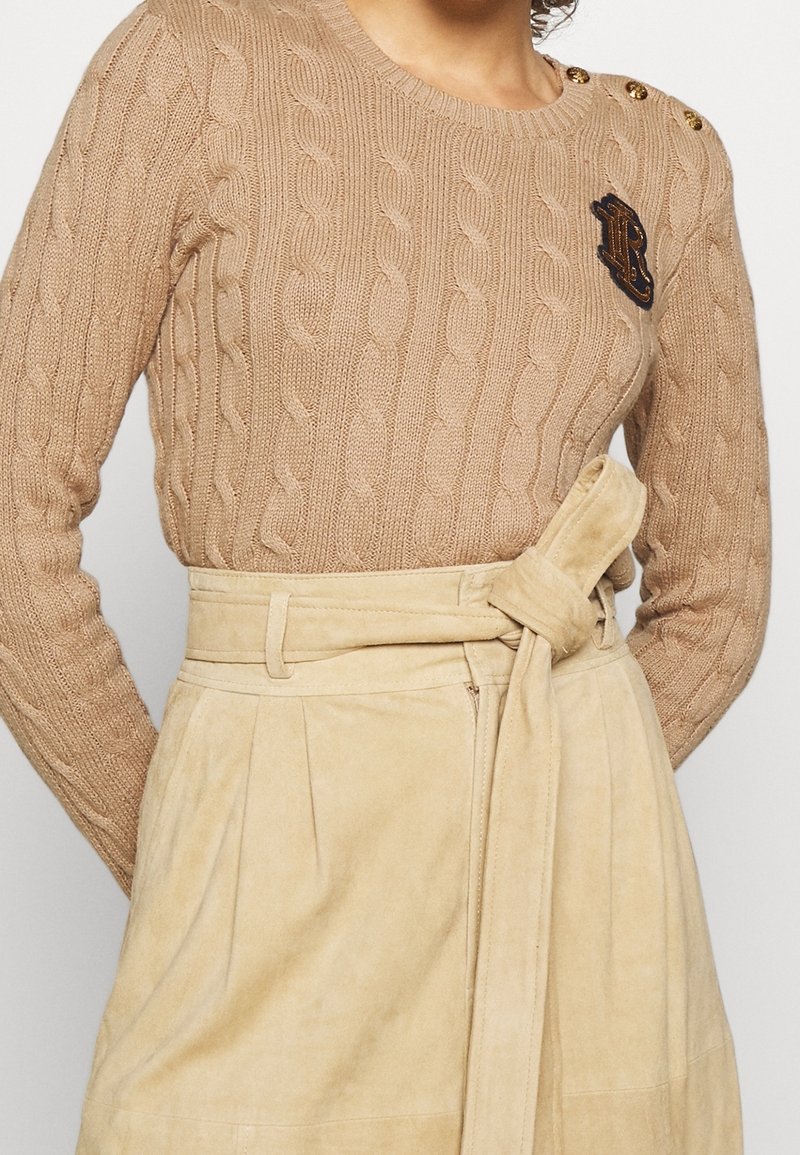 Pull beige en maille torsadée avec des boutons sur l'épaule et un patch brodé foncé, associé à une jupe beige taille haute avec ceinture nouée.