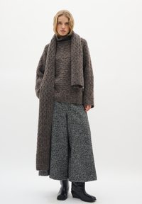 Brauner Strick-Pullover mit Stehkragen, kombiniert mit einem passenden Schal und weiten Hosen mit Hahnentrittmuster, ergänzt durch schwarze Knöchelstiefel.