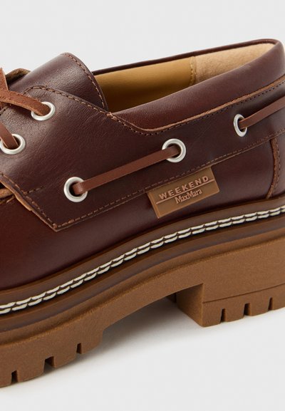 Scarpe da barca in pelle marrone con lacci marroni, occhielli in metallo e suola in gomma testurizzata. Presentano un accento cucito bianco e un'etichetta con il logo.