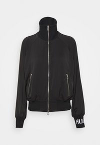 Veste noire zippée avec col montant, deux poches latérales zippées, poignets côtelés et texte "HUG" sur une manchette.