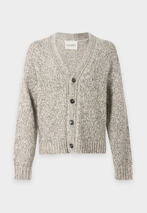 Mand iført en grå knappet cardigan over en brun skjorte, beige viddebukser og sorte sko, stående mod en ensfarvet hvid baggrund.