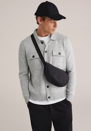 Mand iført sort kasket, lysegrå cardigan med knapper og klaplommer, hvid skjorte, sorte bukser og sort skuldertaske mod neutral baggrund.