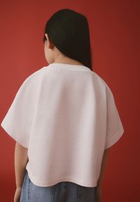 T-shirt en coton blanc avec manches larges et col arrondi, présenté sur un fond rouge. Coupe décontractée et confortable.