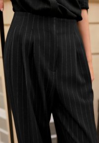 Zwarte pinstripe broeken gemaakt van een textuurstof, met een hoge taille, plooien en zijzakken, afgewerkt met contrasterende witte strepen.
