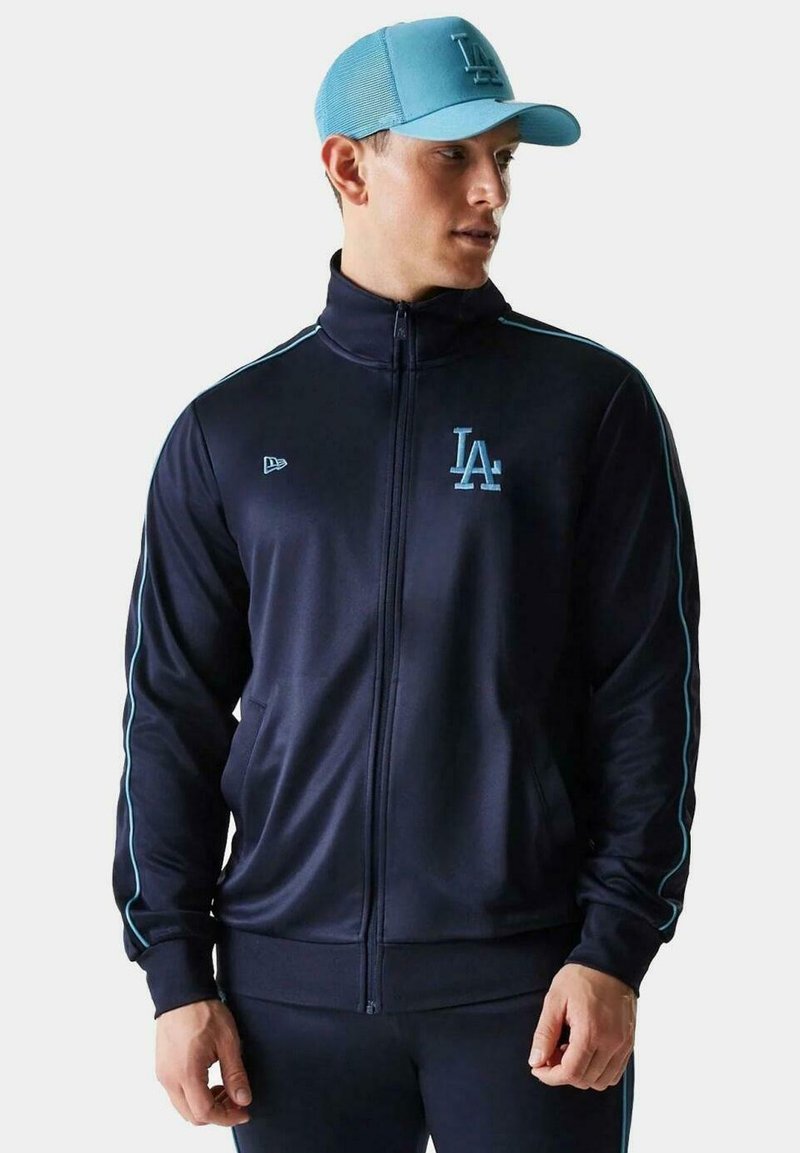 Chaqueta atlética de color navy con detalles en azul claro. Presenta un logo, bolsillos laterales y pantalones oscuros a juego. Gorro azul con diseño estructurado.