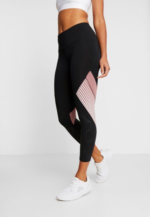 Leggings de sport noirs avec une taille haute, présentant un panneau rose diagonal et un motif rayé sur la jambe inférieure. Portés avec des baskets blanches.